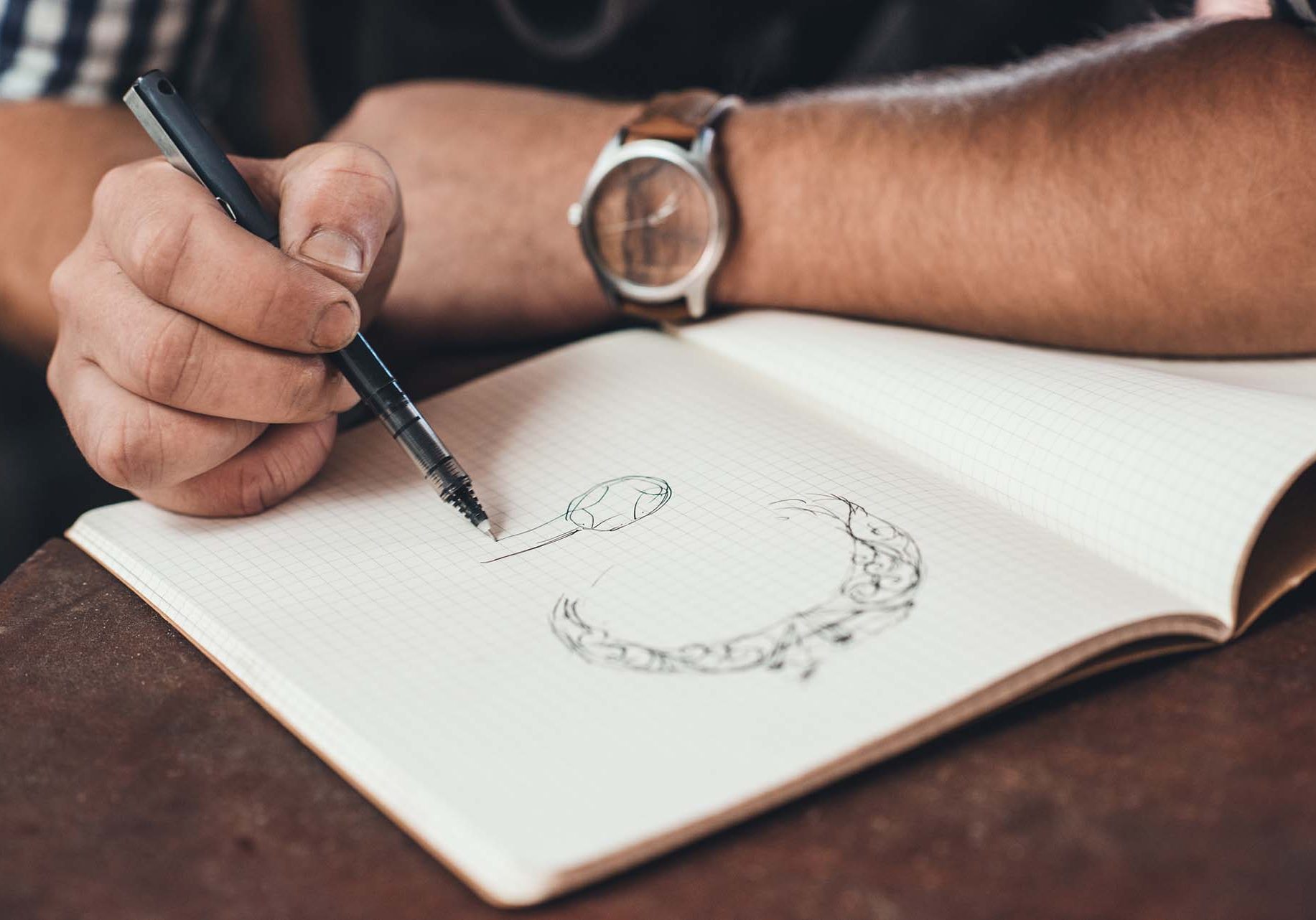 Closeup of a jeweler leaning on a bench sketching out new jewelry designs in a notebook while working in his shop